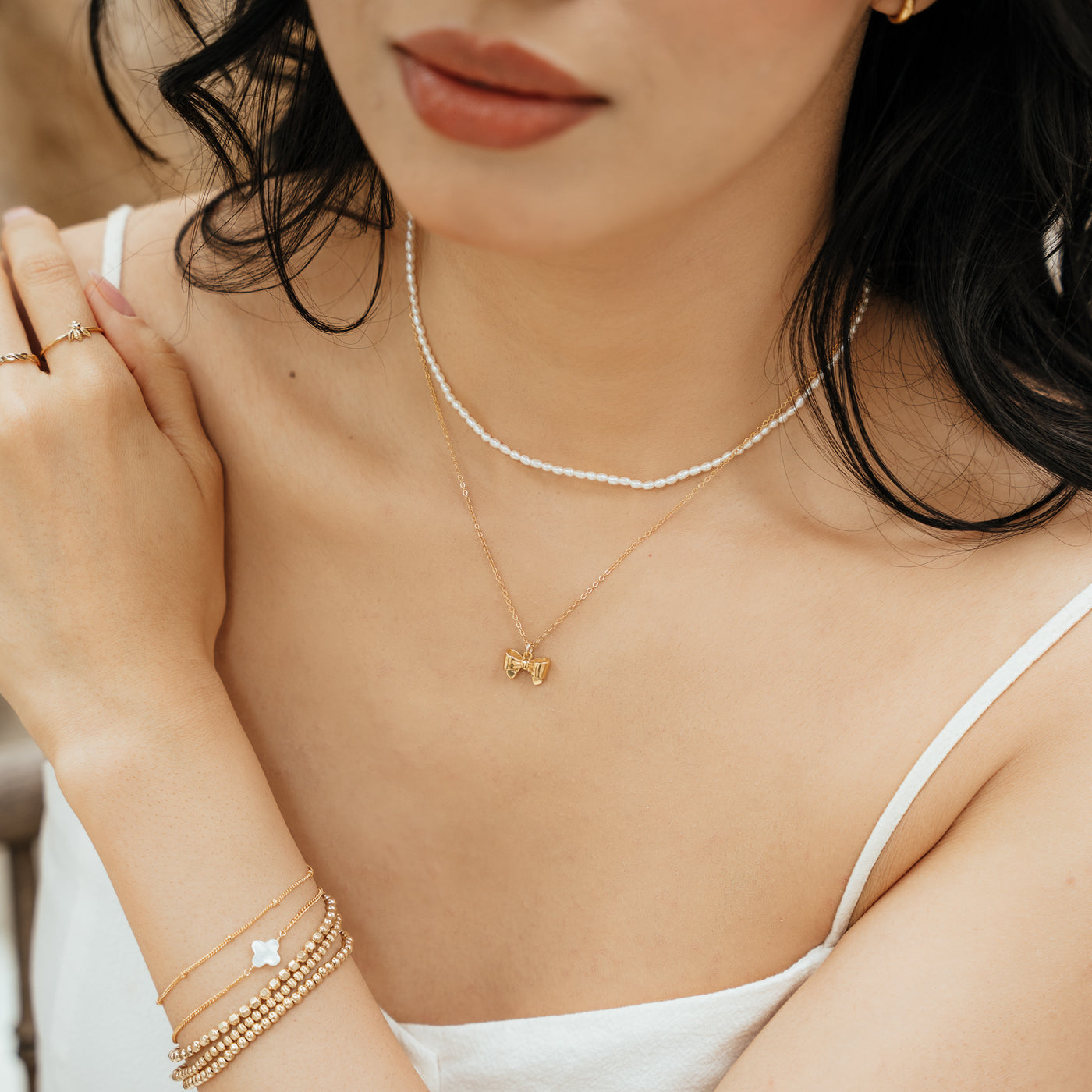 Close-up of a woman’s chest, wearing a Dainty Pearl Strand Necklace, Ribbon Bow Ring, Clover Pearl Bracelet, Satellite Chain Bracelet, and Stretch Bead Bracelets.