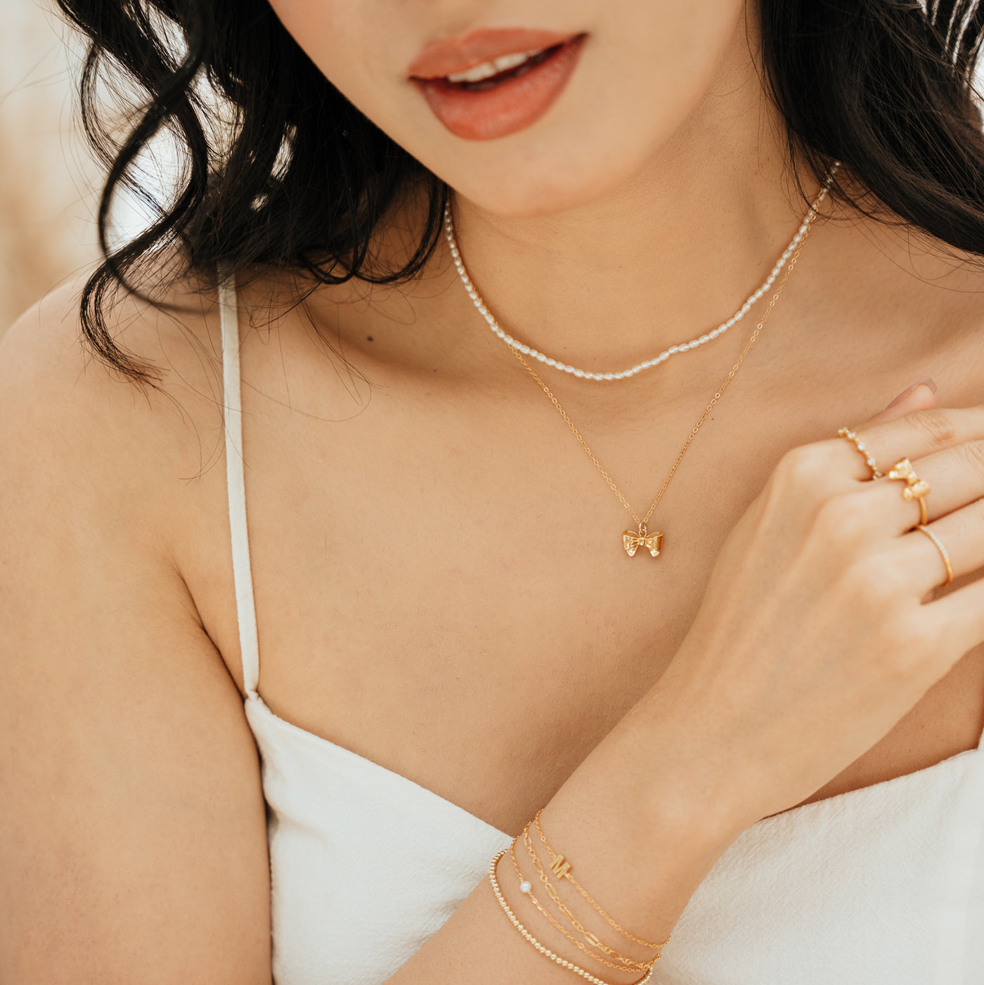 Close-up of a woman’s chest, wearing a Pearl Strand Necklace and Ribbon Bow Necklace. Her right arm crosses her chest with her hand resting on it, showing a Ribbon Bow Ring, Floating CZ Ring, Pavé CZ Ring, and Bracelets.