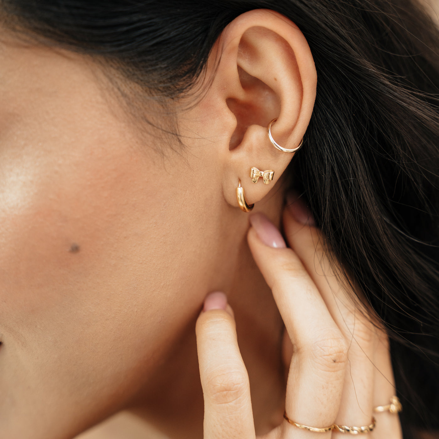Close-up of a woman’s ears wearing the Ribbon Bow Stud Earrings paired with a hoop earring and an ear cuff. Her hand is placed on her neck.