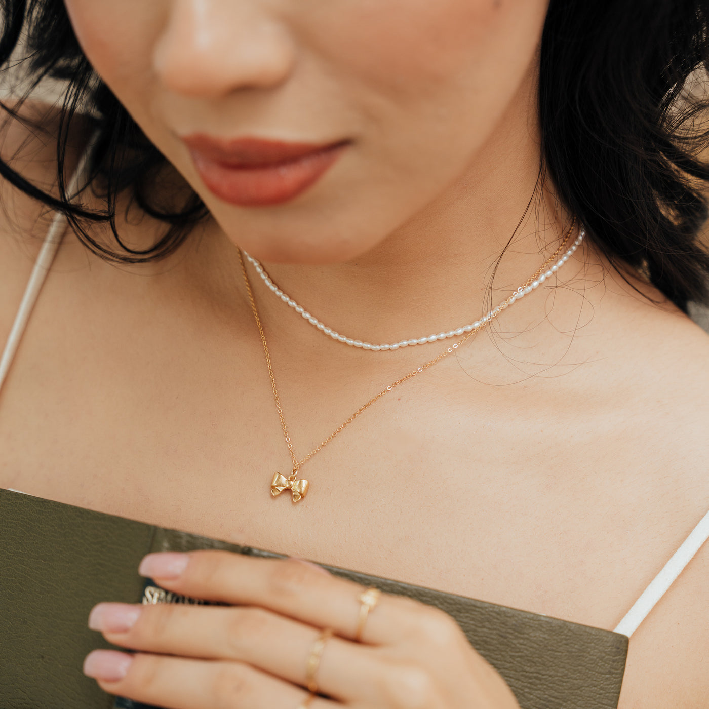 Close-up of a woman’s chest wearing the Dainty Pearl Strand Necklace and the Ribbon Bow Necklace while holding a green book in her hands.