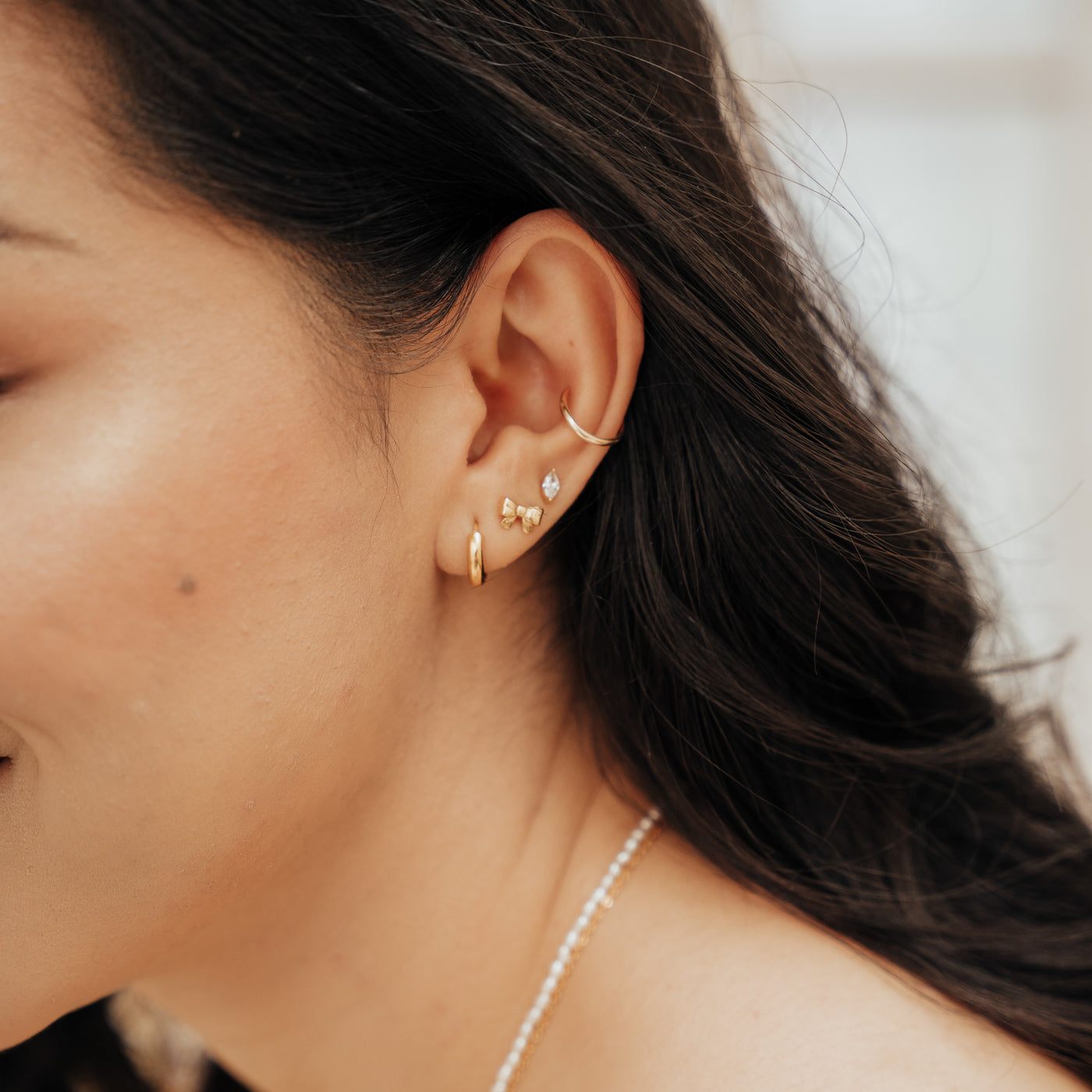 Close-up of a woman’s ear wearing the Ribbon Bow Stud Earrings paired with a hoop earring, a diamond stud earring, and an ear cuff. Around her neck, she is wearing a pearl necklace.