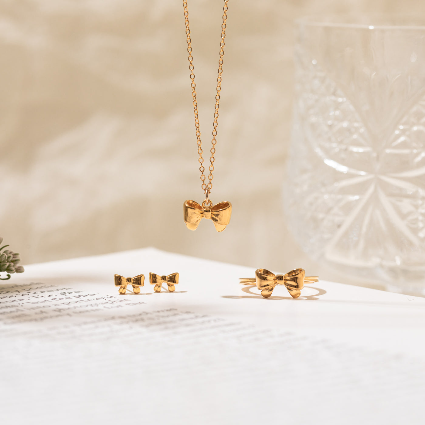 Close-up of the Ribbon Bow Necklace, Ribbon Bow Stud Earrings, and Ribbon Bow Ring placed on a book, with a glass cup beside them.