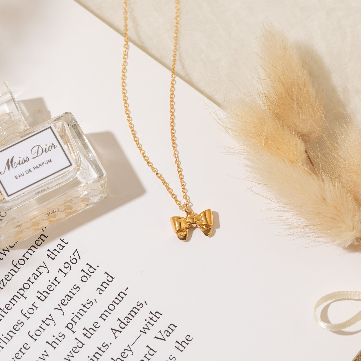 Close-up of the Ribbon Bow Necklace with part of it placed on a book and the rest on soft beige fabric. A perfume bottle and decorative feathers are placed beside it.