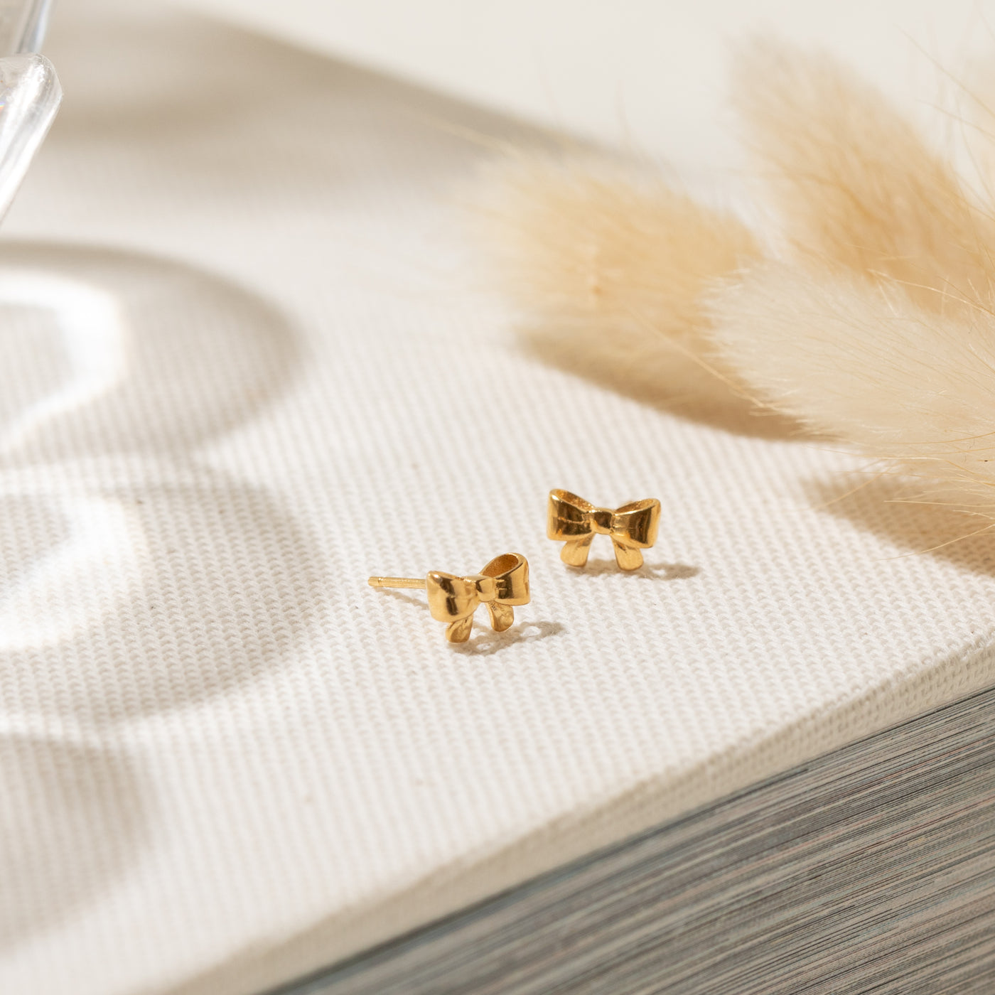 Close-up of the Ribbon Bow Stud Earrings displayed on an off-white surface with decorative feathers beside them.