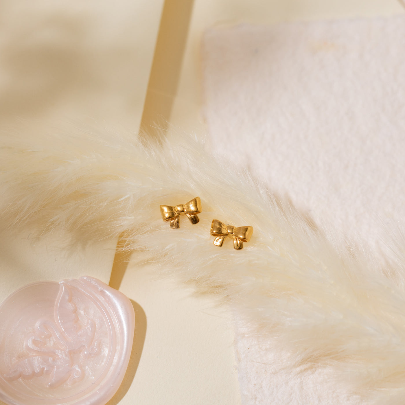 Close-up of the Ribbon Bow Stud Earrings placed on a white feather resting on an envelope with a pink wax seal.