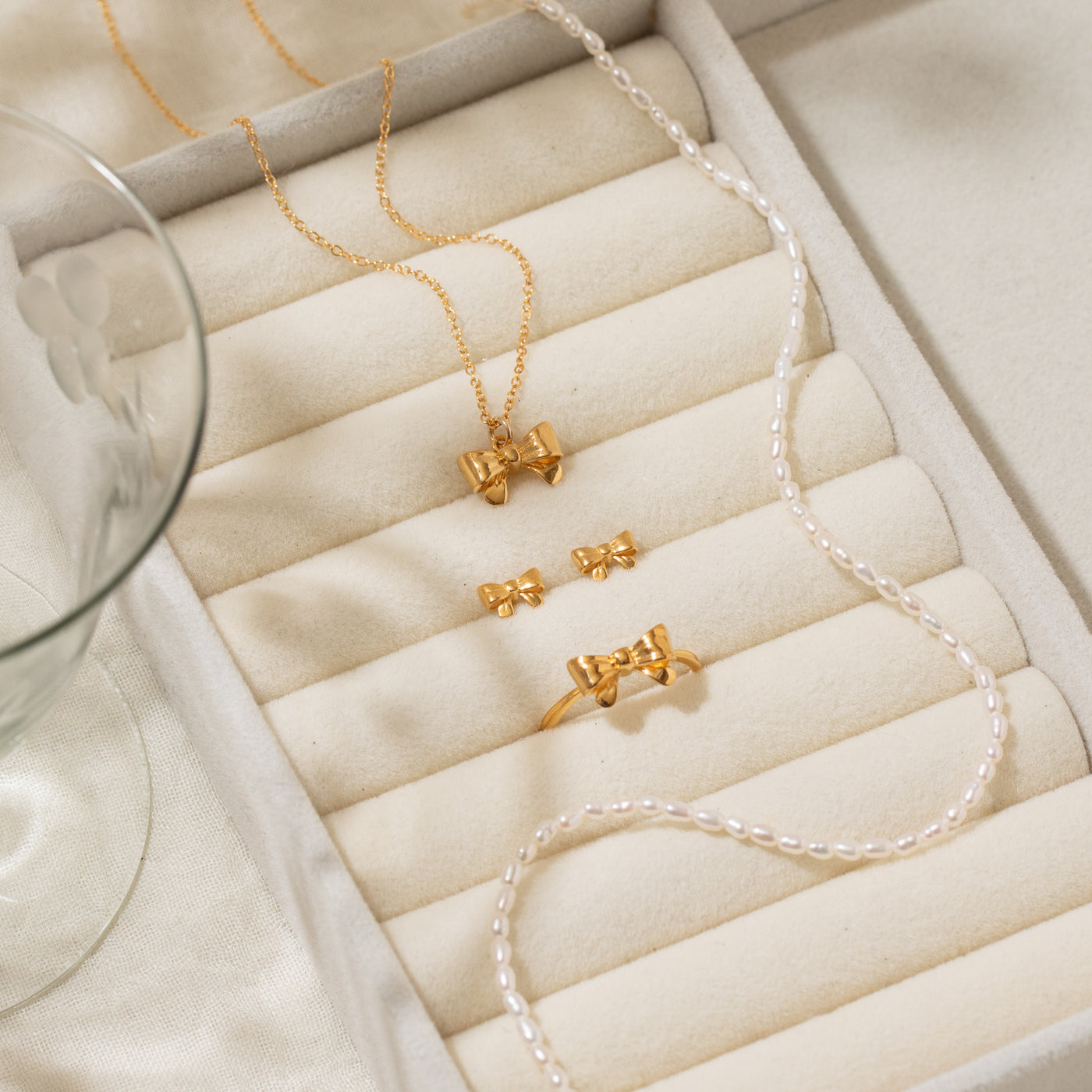 Gold Ribbon Bow Ring, Ribbon Bow Stud Earrings, Ribbon Bow Necklace, and Pearl Strand Necklace displayed on a ring roll jewelry box.