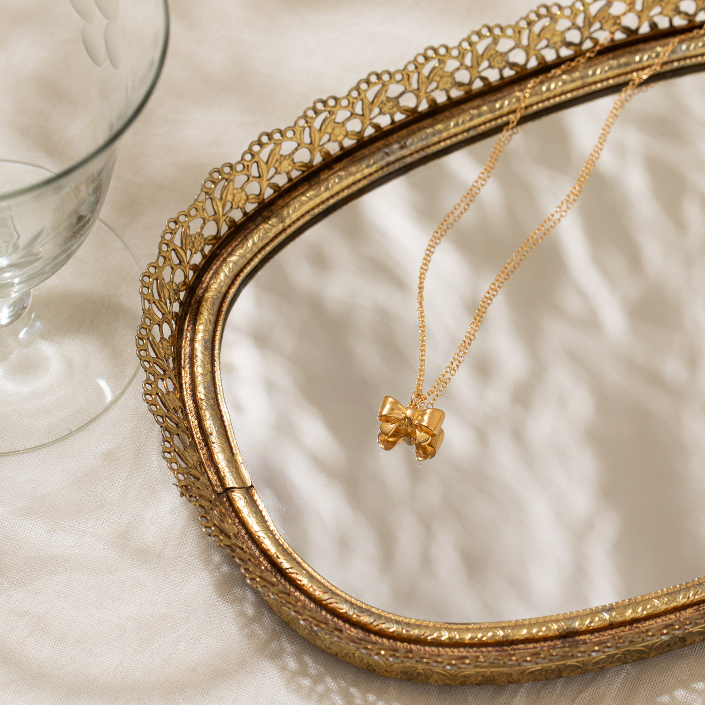 Close-up of the Ribbon Bow Necklace placed on a mirror tray set on soft beige fabric, with a glass cup beside it.