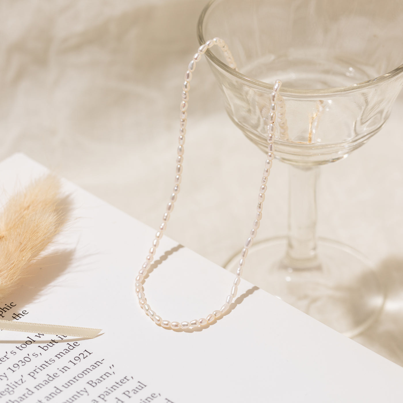 Close-up of the Dainty Pearl Strand Necklace with part of the necklace hanging from a glass cup and resting on a decorative book. A small decorative feather is visible in the corner.