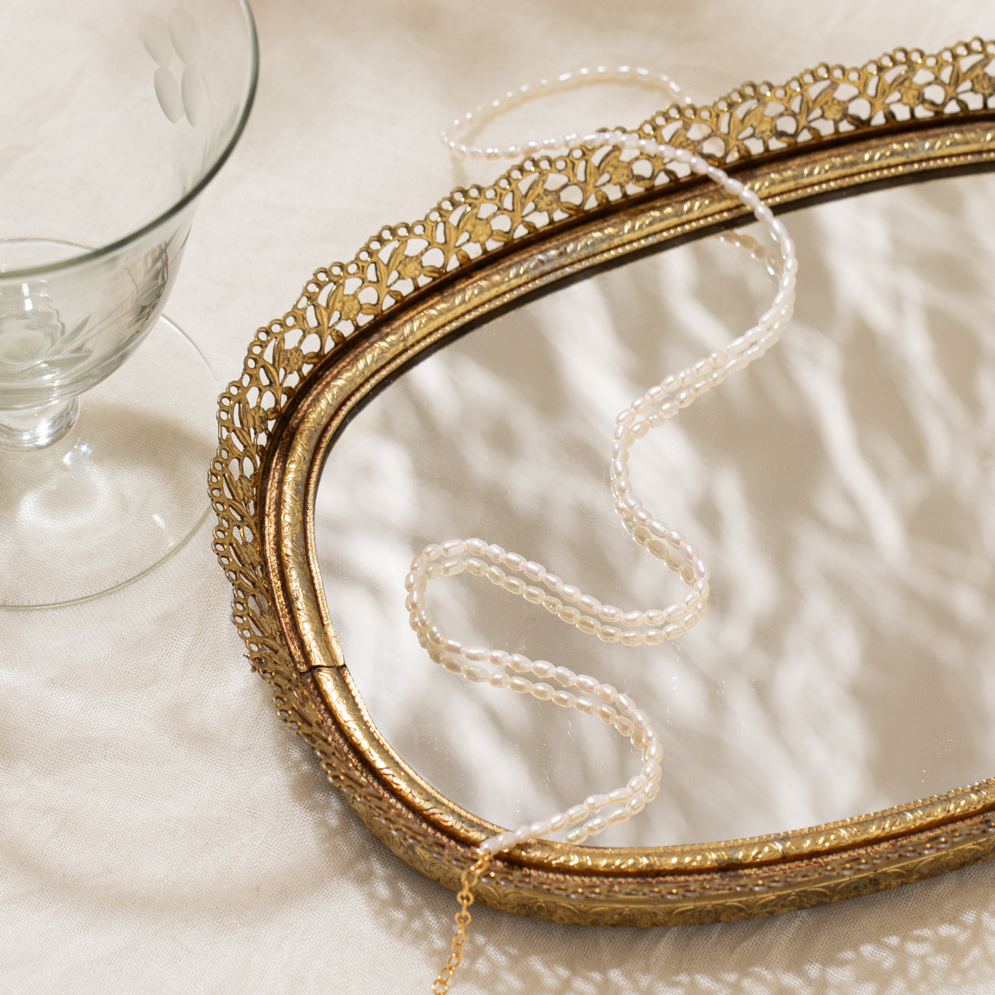 Close-up of the Dainty Pearl Strand Necklace displayed on a mirror tray positioned on soft beige fabric, with a glass cup placed beside it.