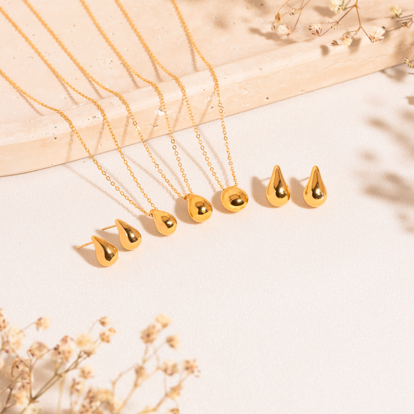Small Large Teardrop Pendant Necklace Several gold pieces lying on white surface, from left to right: dainty teardrop studs, dainty teardrop necklace, teardrop necklace, pebble necklace, and teardrop stud earrings. 