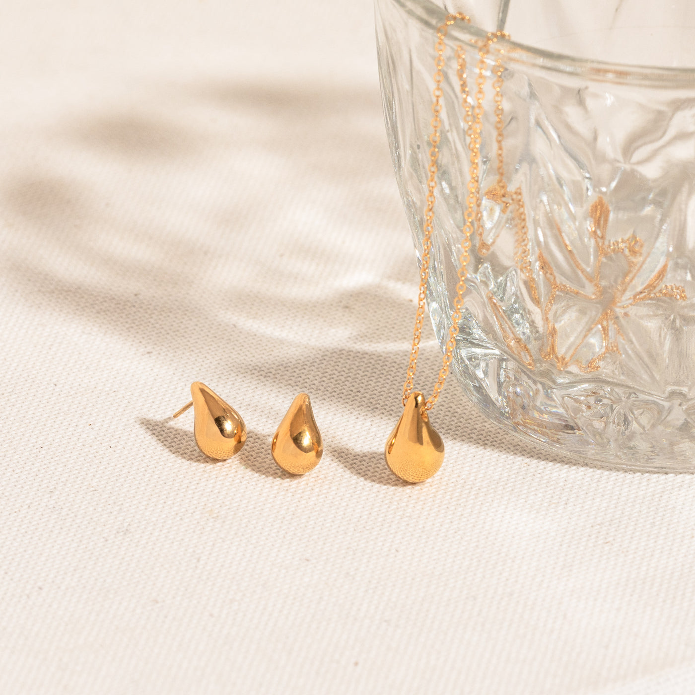 Small Large Teardrop Pendant Necklace gold teardrop stud earrings and teardrop necklace lying on textured white fabric. The teardrop necklace has its chain hanging from a crystal glass