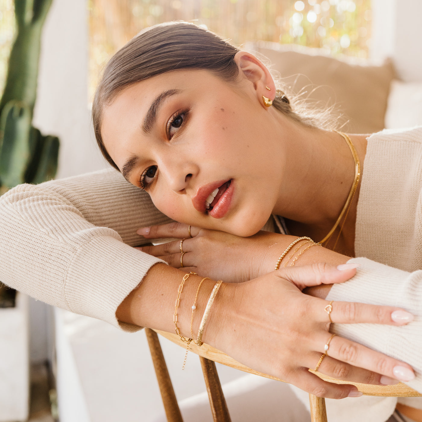 Small Large Teardrop Stud Earrings Woman resting her face on her hand with her arms crossed over the back of a chair, wearing teardrop earrings. On her left wrist, she has a tennis CZ bracelet, a dainty paperclip bracelet, and a heart chain bracelet. On her right wrist, she has a paperclip bracelet, a freshwater pearl bracelet, and a flower cuff bracelet.