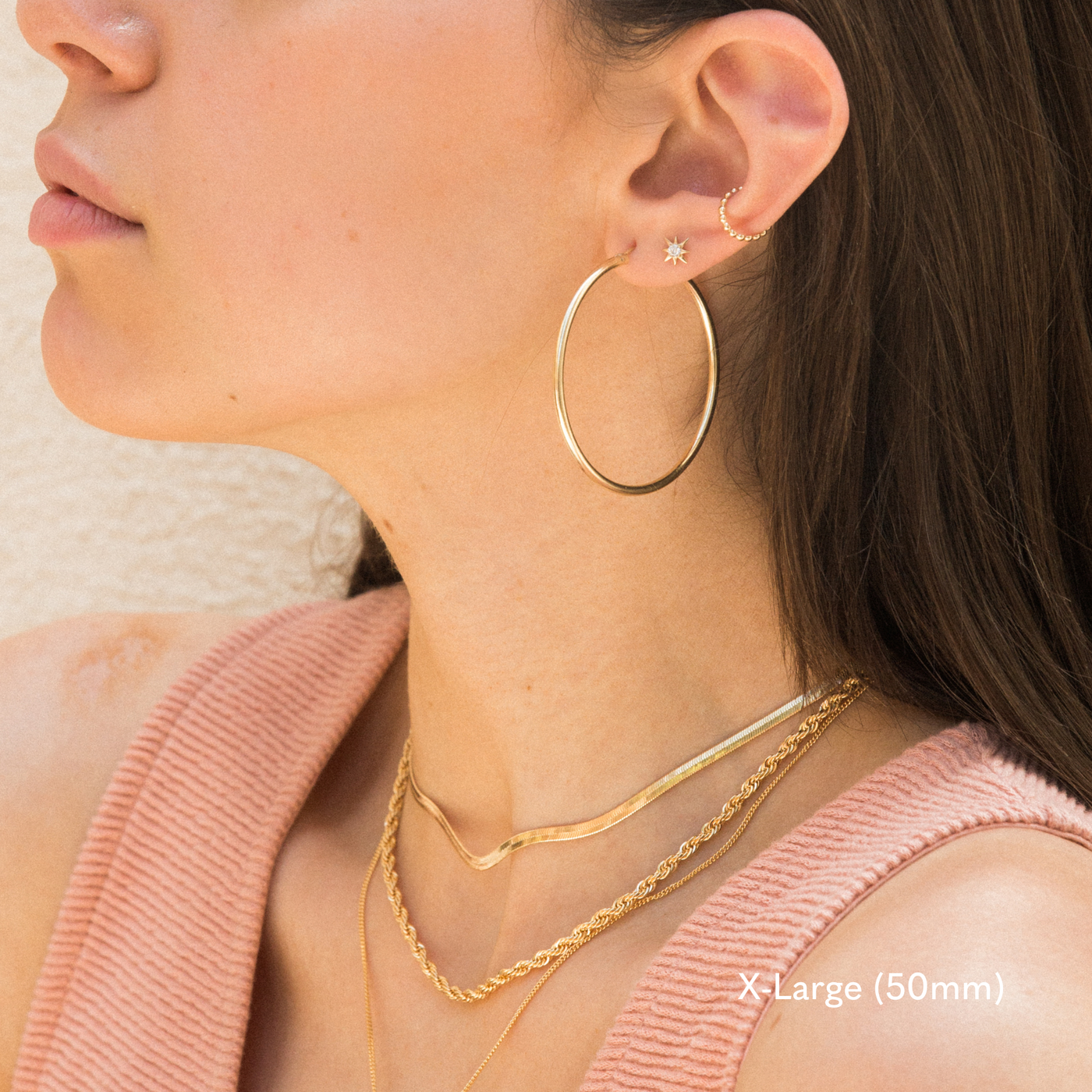 Side profile of a woman wearing an extra large 50mm gold hoop earring, small star stud, and gold ear cuff. She’s dressed in a pink textured tank top and layered gold necklaces. The image is taken in natural light against a light-colored wall.

Small (13mm) Medium (22mm) Large (35mm) X-Large (50mm), Everyday Hoop Earrings by Simple & Dainty Jewelry
