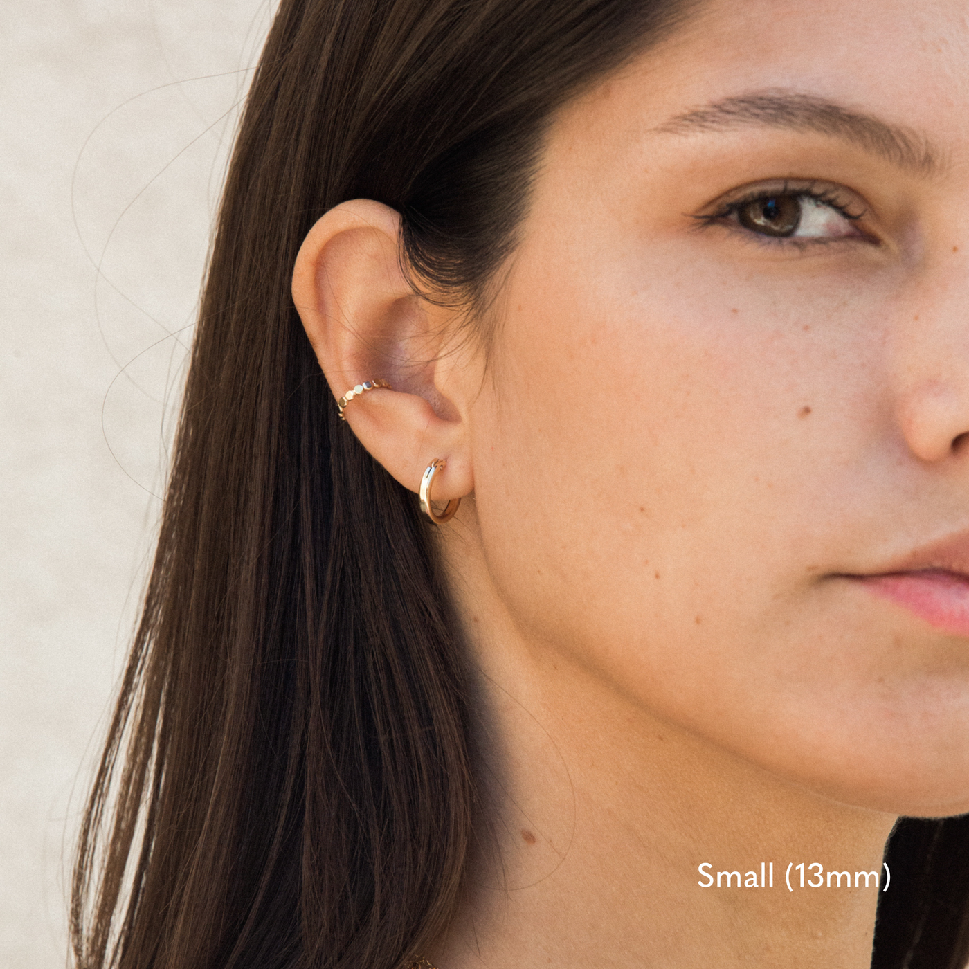 Close-up of a woman wearing a small 13mm gold hoop earring in her left earlobe and a thin gold ear cuff higher on her ear. Her dark hair is tucked behind her ear, and the background is softly lit in neutral tones.

Small (13mm) Medium (22mm) Large (35mm) Everyday Hoop Earrings by Simple & Dainty Jewelry