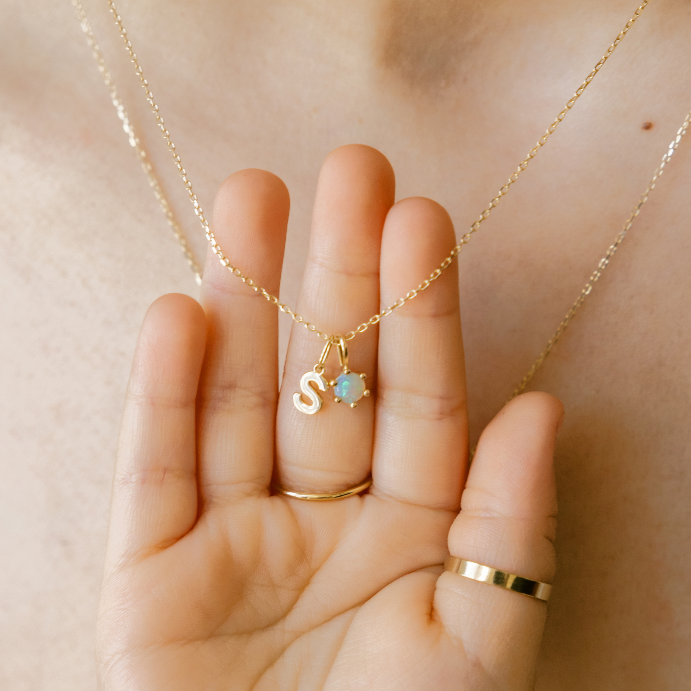 Gold necklace with S and opal held in a child's hand against mother's chest. A B C D E F G H I J K L M N O P Q R S T U V W X Y Z Tiny Hanging Initial Necklace. January - Garnet February - Amethyst March - Aquamarine April - White Topaz April - Diamond May - Emerald June - Pearl June - Moonstone July - Ruby August - Peridot September - Sapphire October - Australian Opal October - Pink Tourmaline November - Citrine December - Tanzanite 14k Birthstone Charm Necklace. 