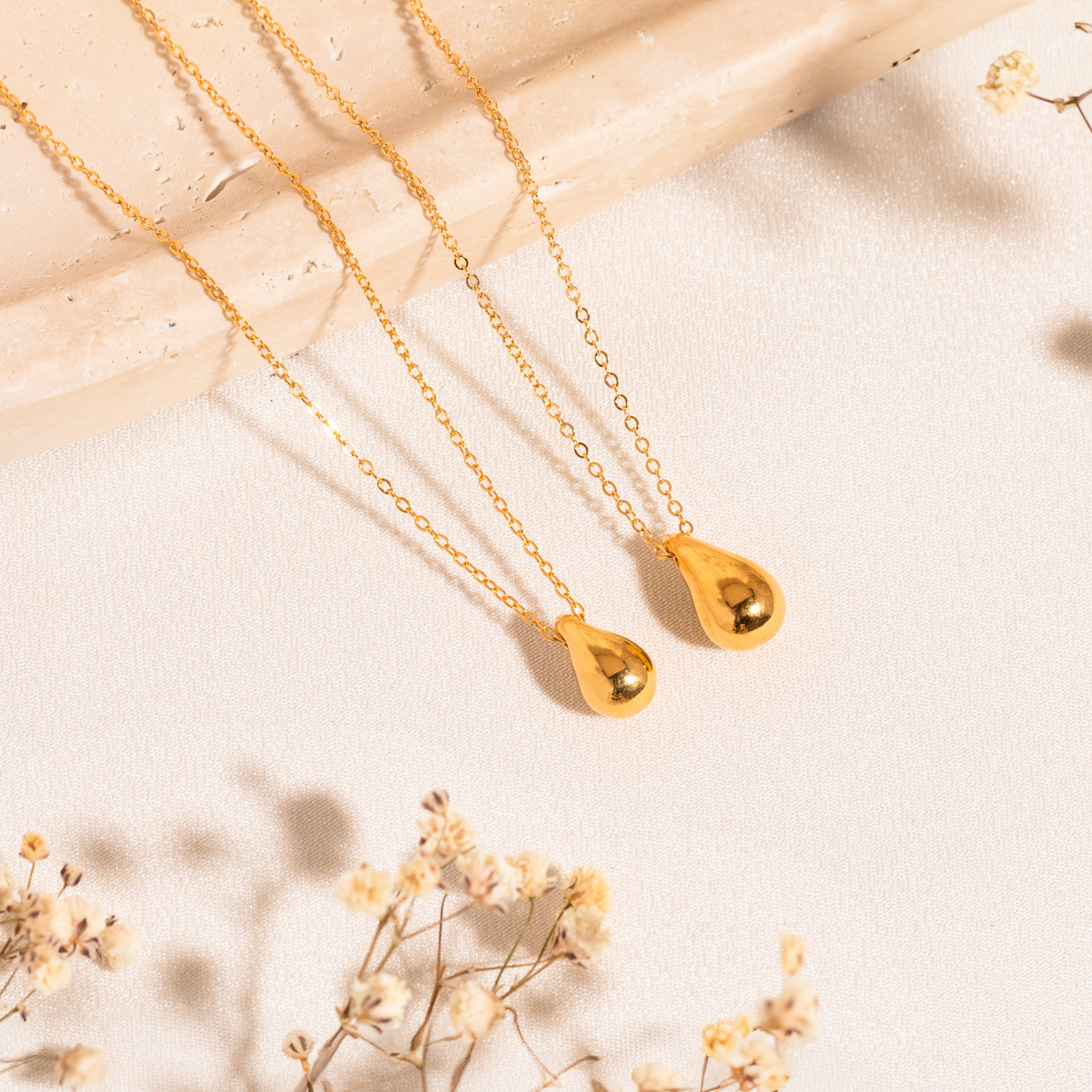 Small Large Teardrop Pendant Necklace Small and large teardrop necklaces lying on a white surface. In the bottom-left corner, there are sprigs of baby's breath, and part of the chains rest on a marble surface.
